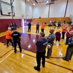 Police officers and volunteers stand in a gymnasium around a basketball court during a community event, with spectators in the bleachers watching.