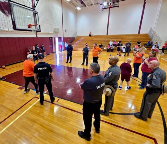 Police officers and volunteers stand in a gymnasium around a basketball court during a community event, with spectators in the bleachers watching.