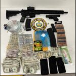 Seized display of illegal items: an AR-style rifle and a handgun with magazines, stacks of cash, cigarette packs, a police badge sticker, several smartphones, and assorted drug paraphernalia on a table.