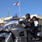 Woman driving a silver touring motorcycle with a child passenger, both wearing helmets, in a parking lot with an American flag in the background.