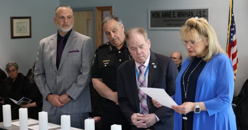 Four adults stand solemnly at a candle-lit table during a formal ceremony; a woman in blue reads a document while others look on, with an american flag in the background.