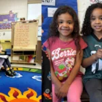 Two young girls sit together in a classroom reading a book; a colorful rug and posters are in the background.