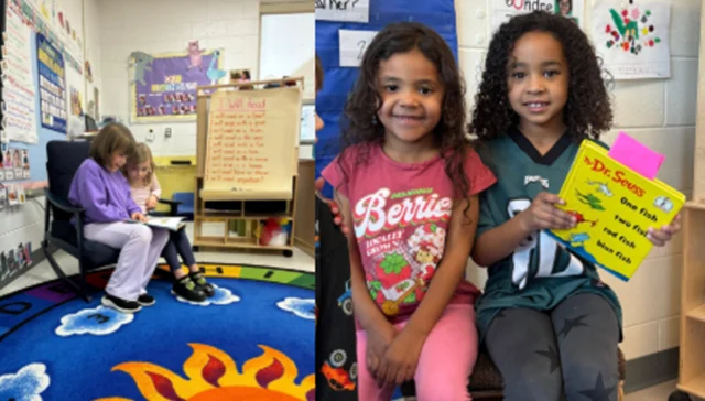 Two young girls sit together in a classroom reading a book; a colorful rug and posters are in the background.