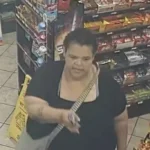Surveillance image of an adult woman in a black top with a striped crossbody bag, standing in a store aisle near snack shelves.