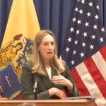 Woman in a blazer speaks at a wooden podium, with U.S. flags and a yellow state flag behind her.
