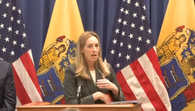 Woman in a blazer speaks at a wooden podium, with u. S. Flags and a yellow state flag behind her.