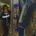 Surveillance image of a man with dreadlocks in a black jacket with a white cross, entering a store.