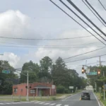 Intersection view with traffic lights overhead, crosswalk, and a red brick building on the left; tangled utility wires and green street signs ahead.