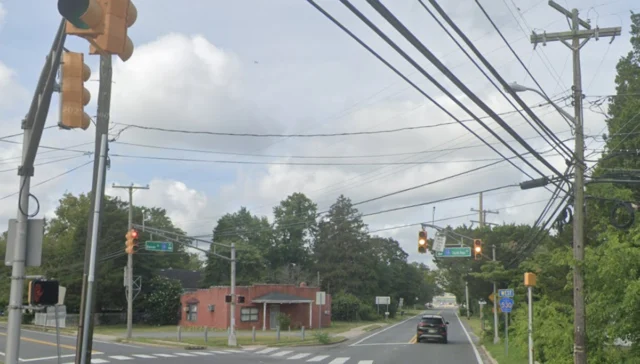 Intersection view with traffic lights overhead, crosswalk, and a red brick building on the left; tangled utility wires and green street signs ahead.
