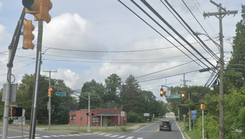 Intersection view with traffic lights overhead, crosswalk, and a red brick building on the left; tangled utility wires and green street signs ahead.
