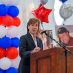 Woman in a dark suit speaks at a wooden podium with a microphone, balloons in red, white, and blue behind her, at a formal event.