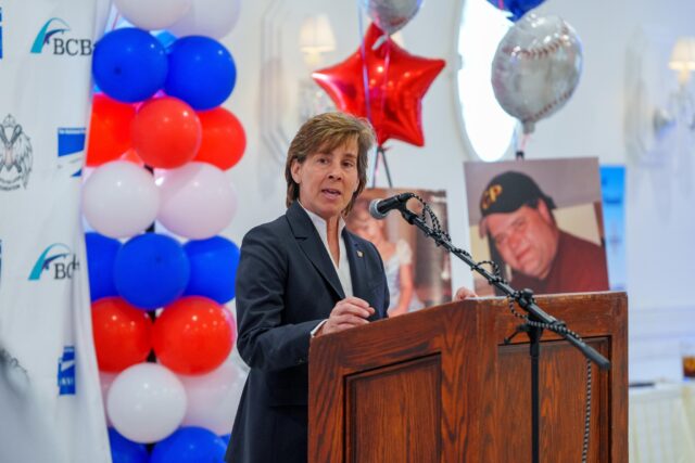 Woman in a dark suit speaks at a wooden podium with a microphone, balloons in red, white, and blue behind her, at a formal event.