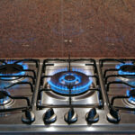 Stainless steel gas stove with four lit burners and blue flames, against a brown granite backsplash.