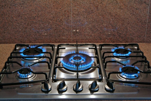 Stainless steel gas stove with four lit burners and blue flames, against a brown granite backsplash.