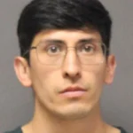 Mugshot of a young man with short dark hair and glasses against a gray backdrop.