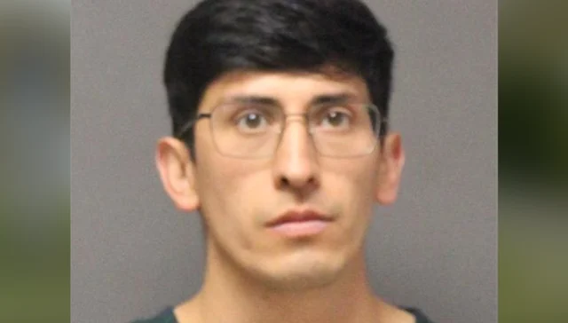 Mugshot of a young man with short dark hair and glasses against a gray backdrop.