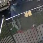 Security camera frame of a man wearing a hat and jacket near a parked car, by a metal fence in a residential area at night/early morning, with some debris on the ground.