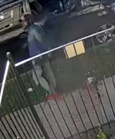 Security camera frame of a man wearing a hat and jacket near a parked car, by a metal fence in a residential area at night/early morning, with some debris on the ground.