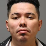 Mugshot of a man facing the camera with a neutral expression, short black hair and light facial stubble against a gray background.