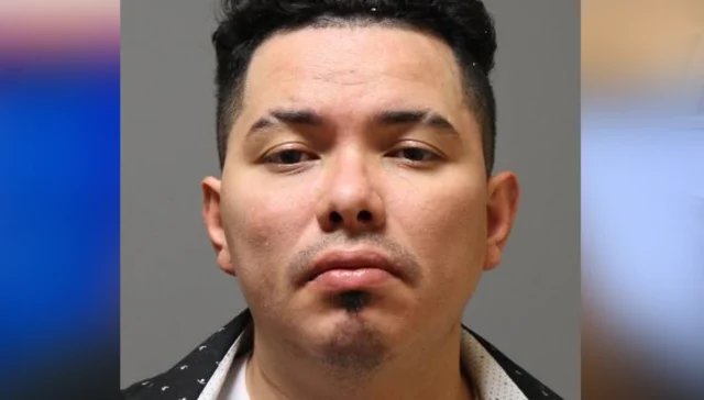 Mugshot of a man facing the camera with a neutral expression, short black hair and light facial stubble against a gray background.