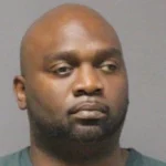 Mugshot of an African-American man with a shaved head and trimmed beard, looking to the left against a gray background.