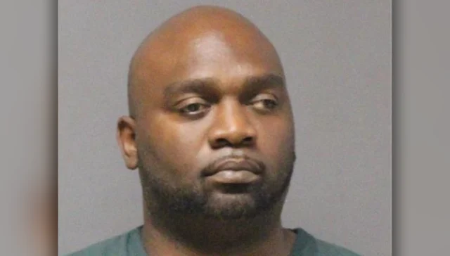 Mugshot of an african-american man with a shaved head and trimmed beard, looking to the left against a gray background.