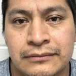 Close-up mugshot of a man with short dark hair and light stubble, facing the camera against a white wall background.
