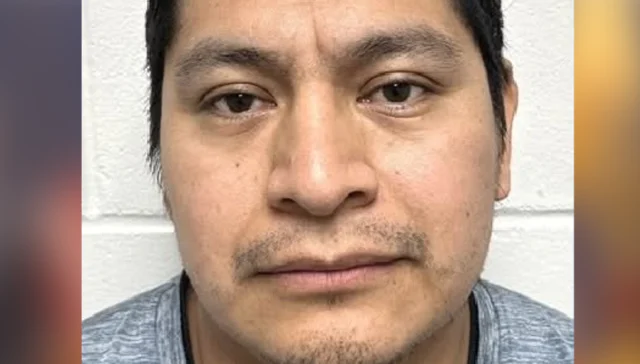 Close-up mugshot of a man with short dark hair and light stubble, facing the camera against a white wall background.