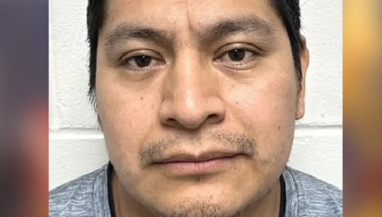 Close-up mugshot of a man with short dark hair and light stubble, facing the camera against a white wall background.