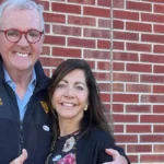Older man in a navy jacket with red glasses, text 'Governor Phil Murphy,' embracing a woman against a red brick wall.