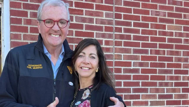Older man in a navy jacket with red glasses, text 'Governor Phil Murphy,' embracing a woman against a red brick wall.