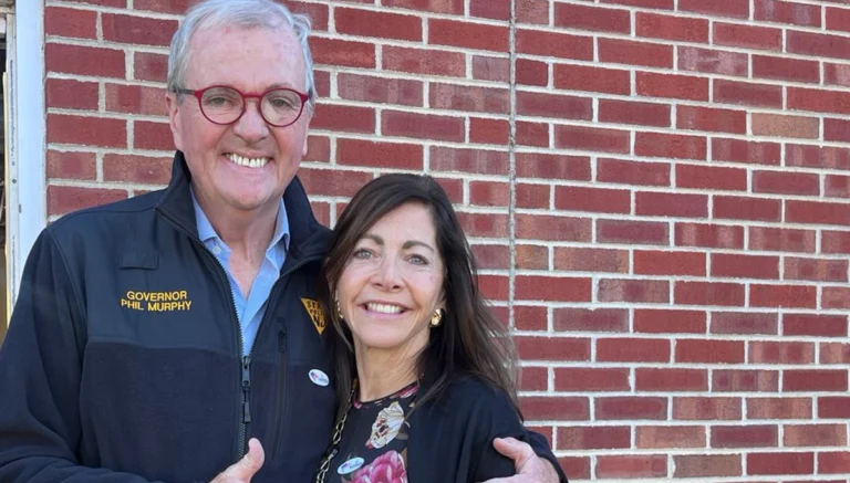 Older man in a navy jacket with red glasses, text 'Governor Phil Murphy,' embracing a woman against a red brick wall.