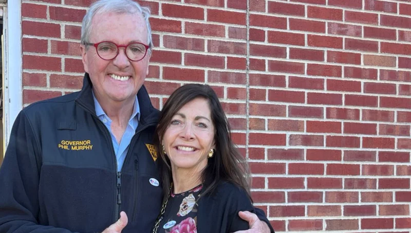 Older man in a navy jacket with red glasses, text 'governor phil murphy,' embracing a woman against a red brick wall.