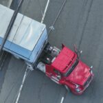 Overhead view of a red semi-truck attached to a blue trailer stopped on a cracked road, with power lines crossing above the vehicles.