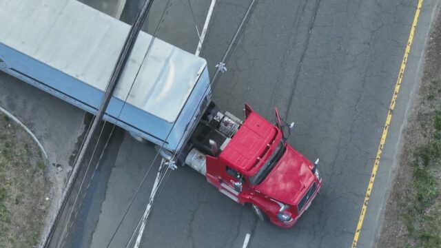 Overhead view of a red semi-truck attached to a blue trailer stopped on a cracked road, with power lines crossing above the vehicles.