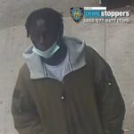 Surveillance image of a man with short dreadlocks wearing a brown jacket with a gray hood and a light mask, standing outdoors; NYPD Crime Stoppers logo in the corner indicating a wanted-suspect photo.