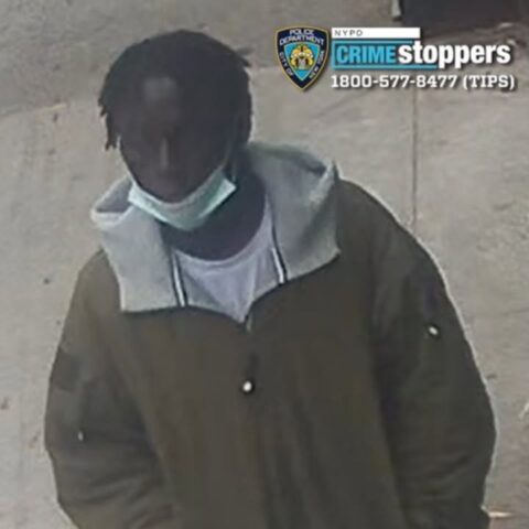 Surveillance image of a man with short dreadlocks wearing a brown jacket with a gray hood and a light mask, standing outdoors; nypd crime stoppers logo in the corner indicating a wanted-suspect photo.
