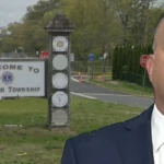 Man in a navy suit and red tie stands beside a 'Welcome to Jackson Township' sign at a road entrance.