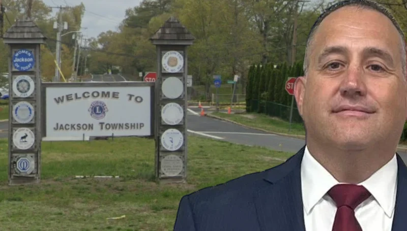 Man in a navy suit and red tie stands beside a 'welcome to jackson township' sign at a road entrance.