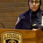 Police officer speaking at a podium with a police badge emblem visible on the front panel.