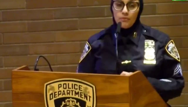 Police officer speaking at a podium with a police badge emblem visible on the front panel.