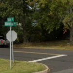 Intersection of Beam Ave and Manhattan St with a stop sign and a white pickup parked by trees in a residential area.