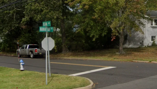 Intersection of beam ave and manhattan st with a stop sign and a white pickup parked by trees in a residential area.