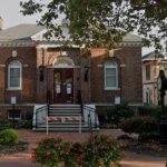 Brick town hall with red doors, arched windows, and a central staircase framed by black railings and a construction barrier.