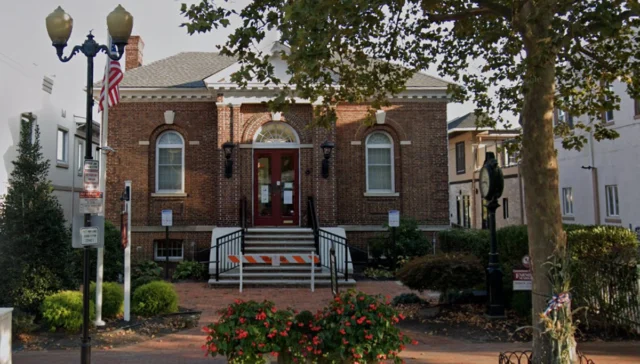 Brick town hall with red doors, arched windows, and a central staircase framed by black railings and a construction barrier.