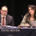 Panelists at a formal meeting: man on left (Michael Citta) speaks into a microphone, woman on right (President Ashley Lamb) writes notes, with 'TOMS RIVER' on the tablecloth.