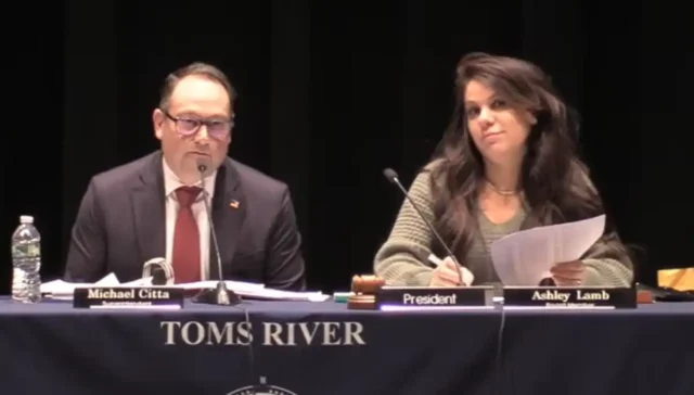 Panelists at a formal meeting: man on left (michael citta) speaks into a microphone, woman on right (president ashley lamb) writes notes, with 'toms river' on the tablecloth.