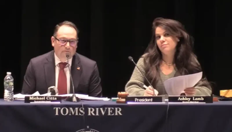 Panelists at a formal meeting: man on left (michael citta) speaks into a microphone, woman on right (president ashley lamb) writes notes, with 'toms river' on the tablecloth.