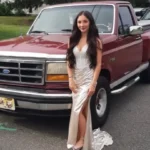 Young woman in a champagne gown with a high slit posing beside a maroon Ford pickup truck in a parking lot.