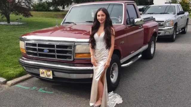 Young woman in a champagne gown with a high slit posing beside a maroon ford pickup truck in a parking lot.
